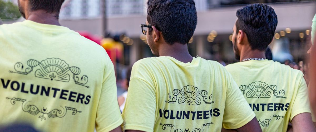 Three volunteers in yellow VOLUNTEERS T-shirts standing with their backs to the camera at a community event