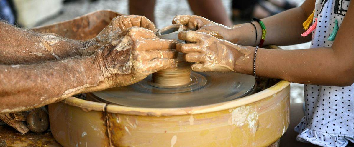 An adult's hands beside a child's hands, shaping a pot together on a potter's wheel